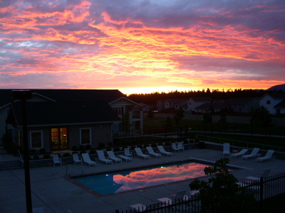 [The sun is setting behind the buildings and evergreens and it appears the clouds are on fire from the bottom. This pink and white cloud color is reflected in the swimming pool which has white chairs around it on three sides.]